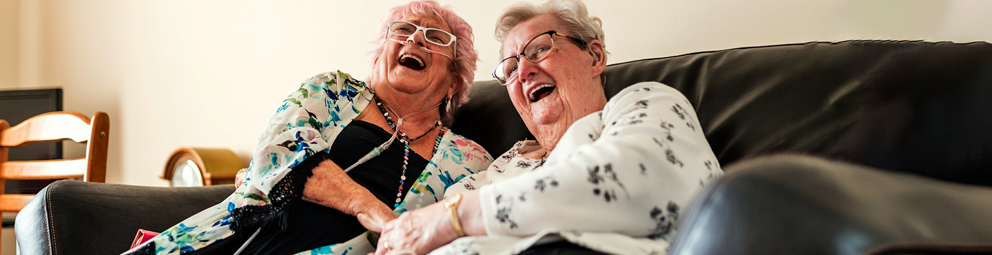 Two older adult women laughing on a couch. One of them has a nasal cannula for oxygen